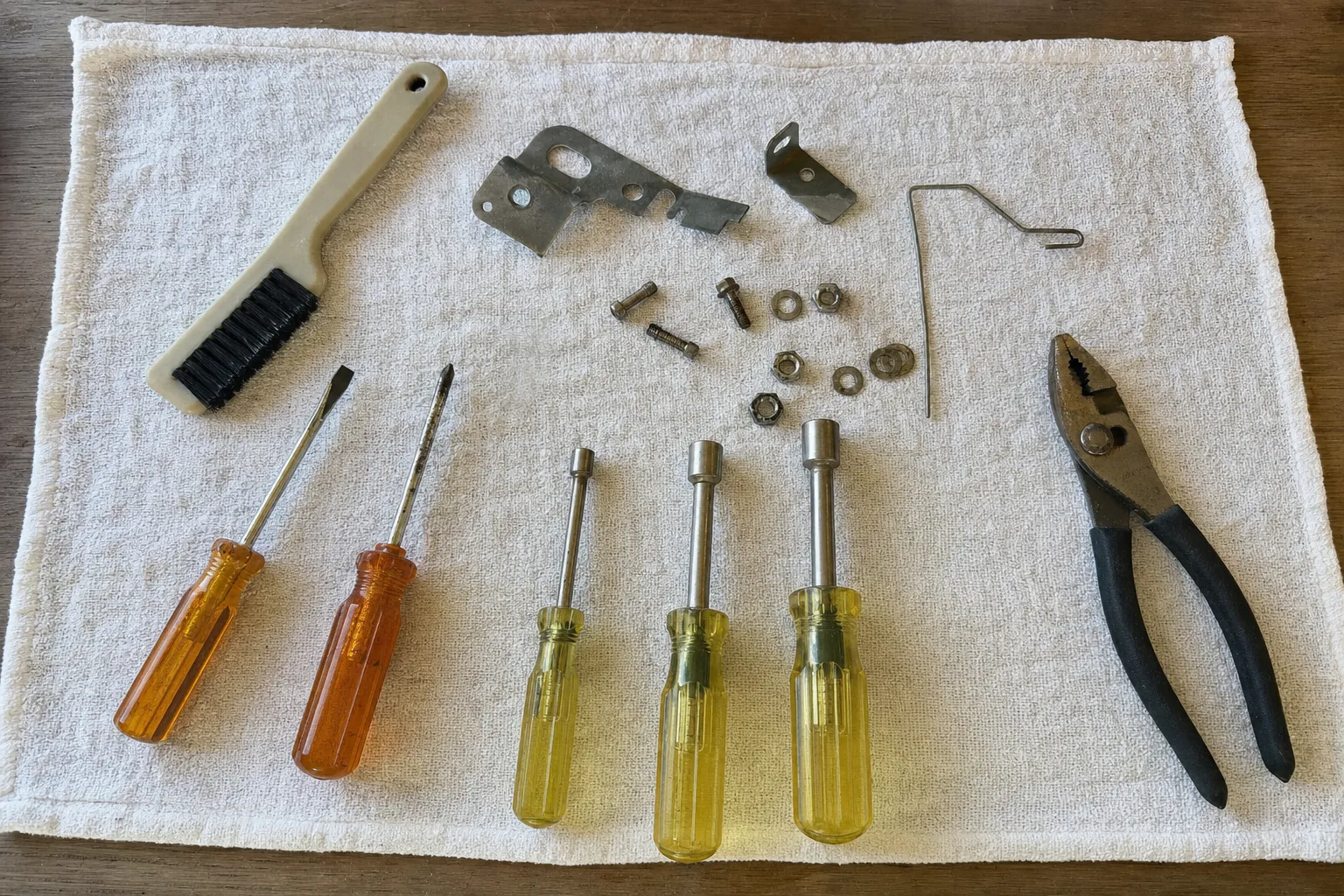 Two screwdrivers, three yellow nut drivers, slip-joint pliers and a brush on a white towel with two stamped brackets, a bent wire rod, nuts and washers